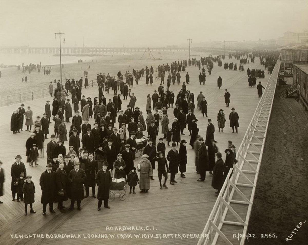 A large crowd of men, women, and children in early 20th-century clothing stroll along a wide wooden boardwalk by the beach, with the ocean and piers visible in the background.