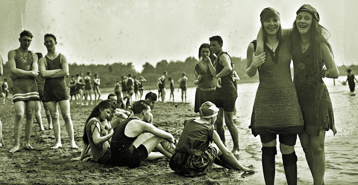 Vintage photo of people at a beach; groups of men and women in early 20th-century swimwear are standing, talking, or sitting on the sand. Two women in the foreground smile at the camera, giving thumbs up.