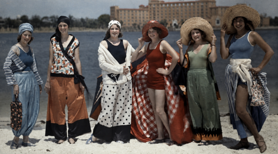 Six women stand on a beach in colorful, vintage 1920s swimwear and wide-brimmed hats, smiling at the camera, with water and a large hotel building visible in the background.
