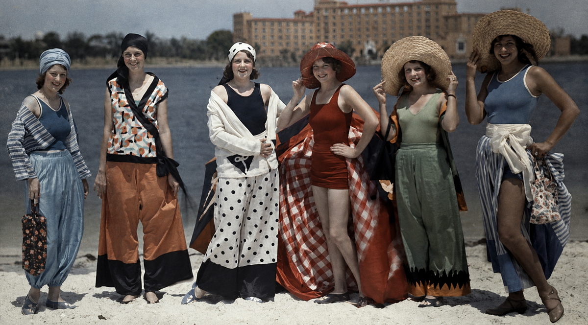 Six women stand on a beach in colorful, vintage 1920s swimwear and wide-brimmed hats, smiling at the camera, with water and a large hotel building visible in the background.