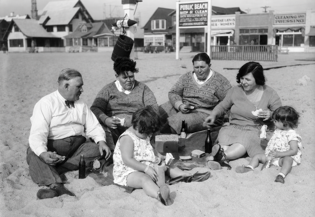 A black-and-white photo shows three adults and three children sitting on the sand having a picnic at the beach, with old-fashioned buildings and signs in the background. The adults appear to be sharing food and drinks.