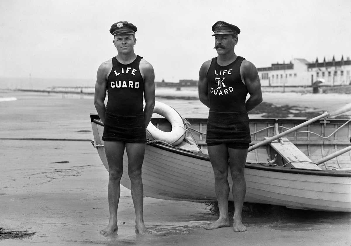 Two male lifeguards in vintage uniforms stand barefoot on the beach in front of a lifeboat, with buildings and the shoreline visible in the background.