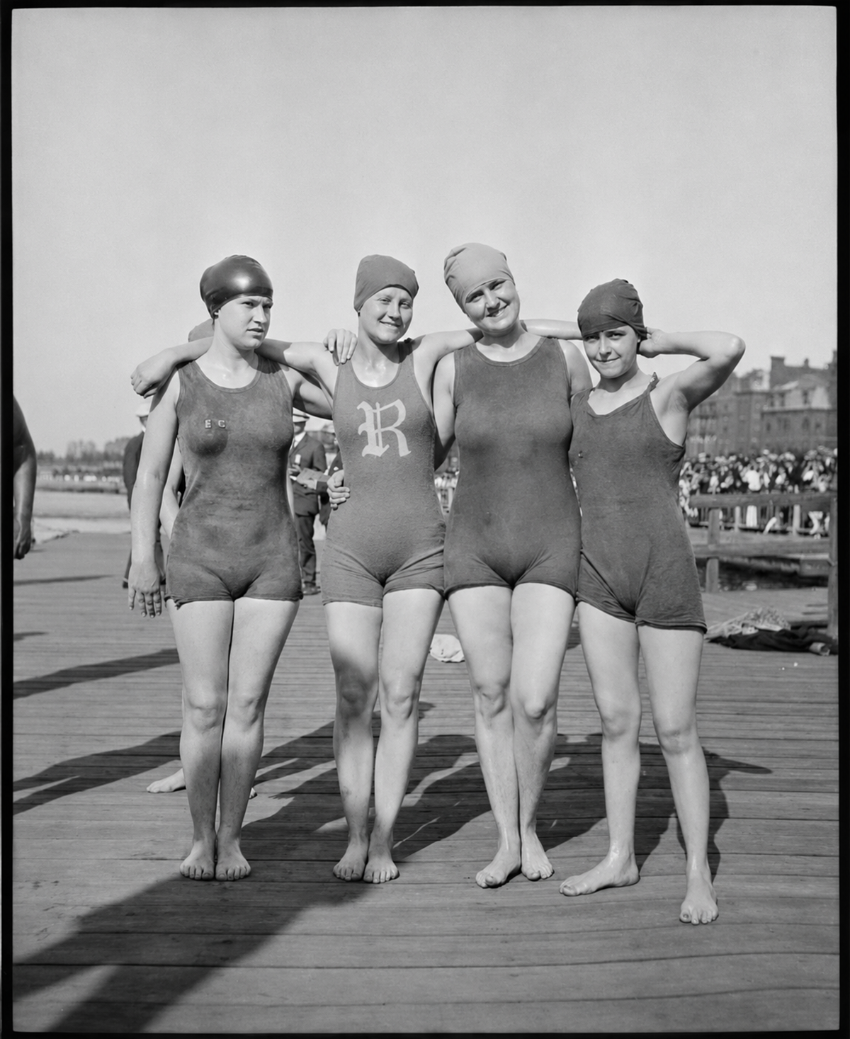 Four women in vintage swimsuits and swim caps stand arm in arm on a wooden boardwalk by the beach, smiling for the camera with a crowd and buildings in the background.