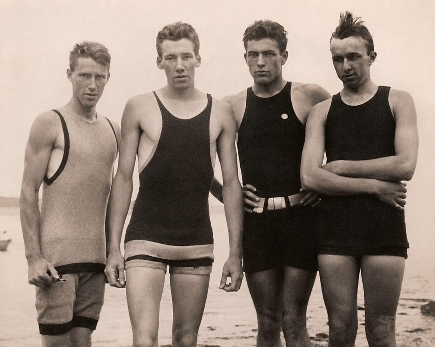 Four young men in vintage swimsuits stand together on a beach, posing for the camera. The photo is black and white, suggesting it was taken in the early 20th century.