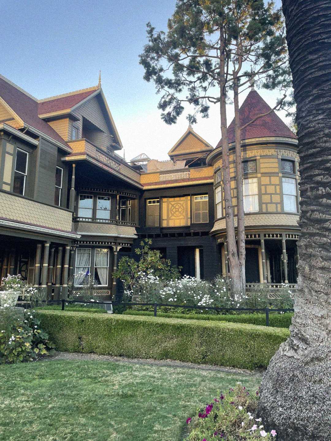 A large, ornate Victorian house with towers and intricate woodwork stands behind a neatly trimmed lawn and garden, framed by tall trees under a clear sky.