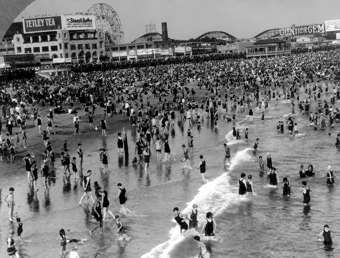 A crowded beach scene from the early 20th century shows hundreds of people wading and swimming in the water, with vintage buildings, advertisements, and a roller coaster visible in the background.