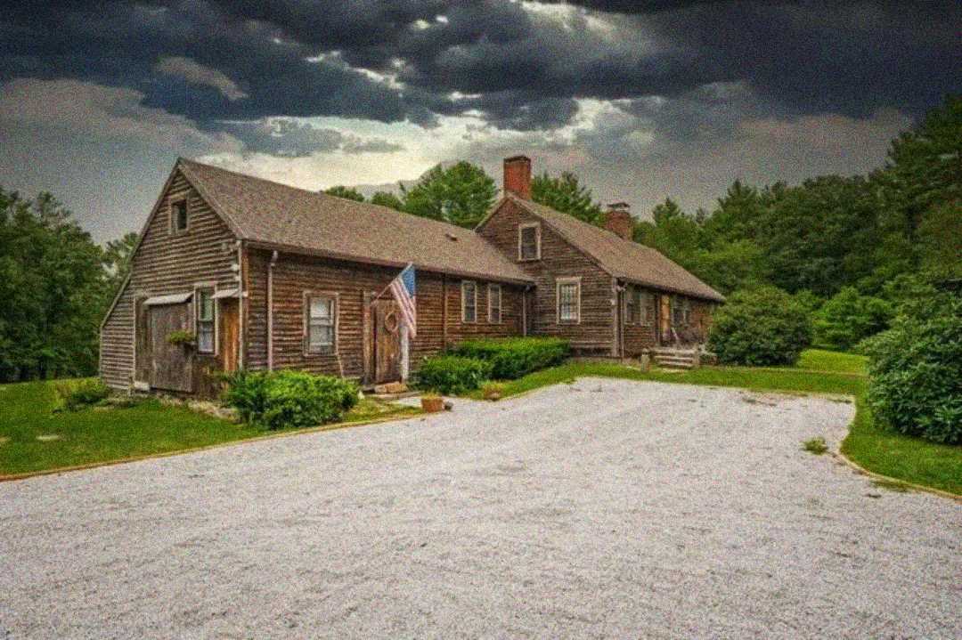 A rustic wooden house with an American flag by the door sits amid greenery under a cloudy, dark sky, with a large gravel driveway in front.