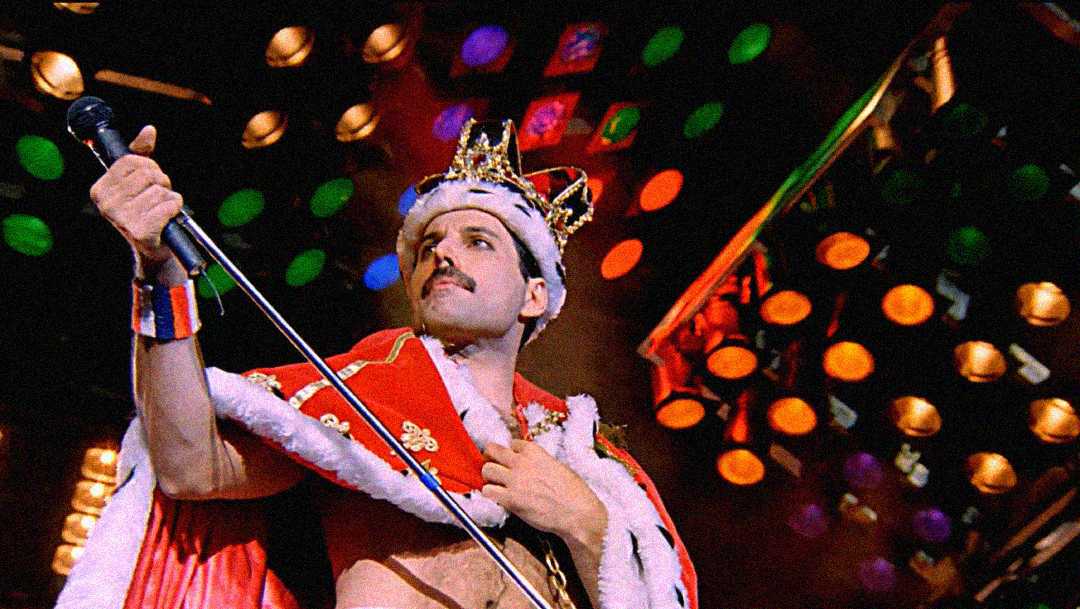 A performer wearing a crown and a red cape with white trim holds a microphone stand on stage, with colorful lights in the background.