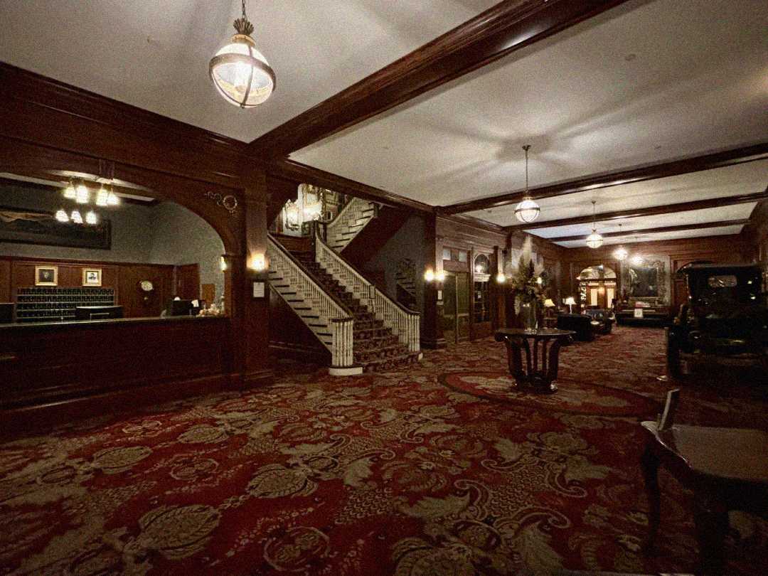 A grand hotel lobby with ornate red carpet, dark wood paneling, chandeliers, a large staircase, a reception desk, vintage furniture, and an antique car near the entrance. The atmosphere feels historic and elegant.