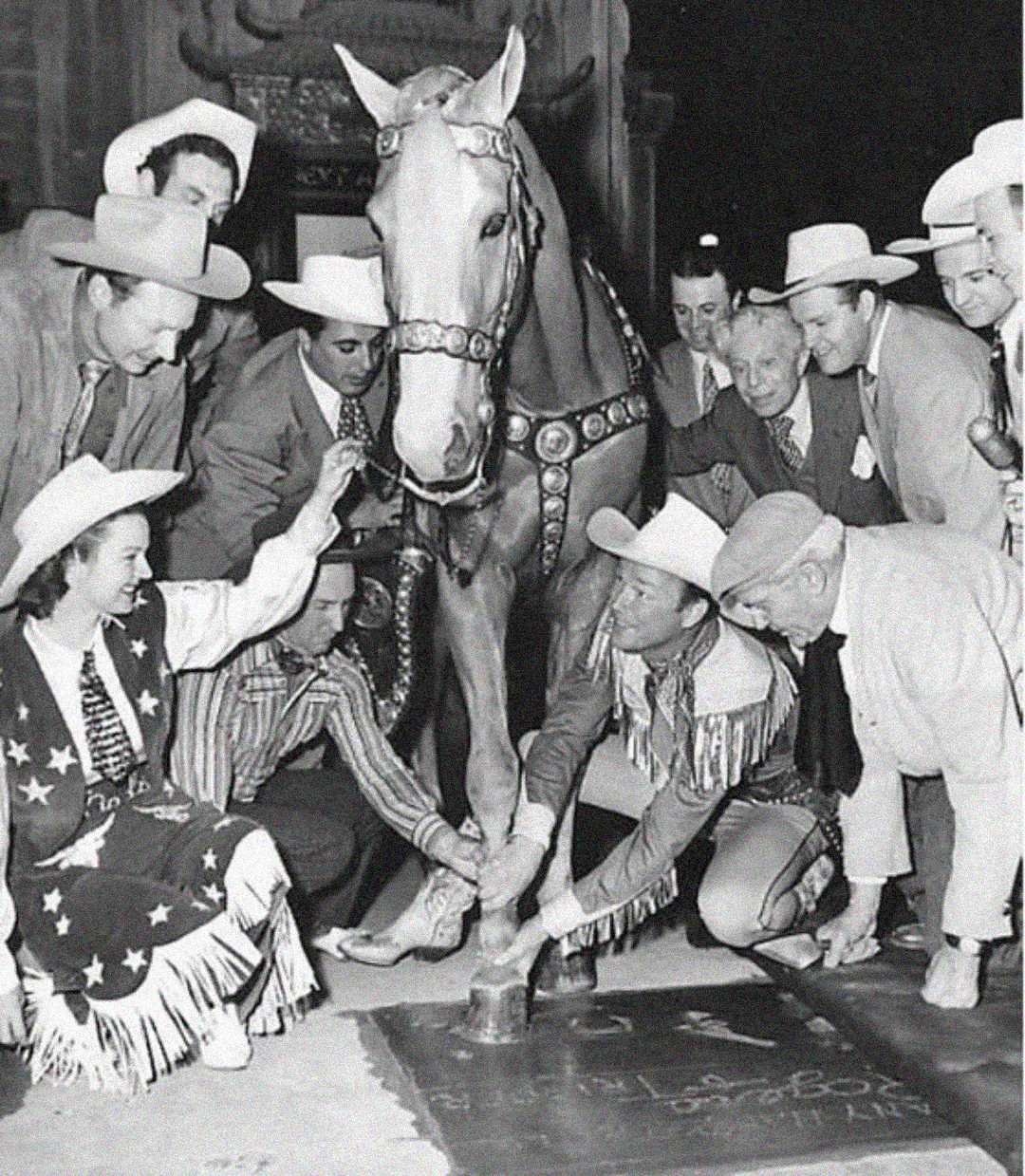 A group of people in cowboy hats and western attire surround a horse, guiding its hoof to make an imprint in wet cement on the ground. The scene appears celebratory and ceremonial.