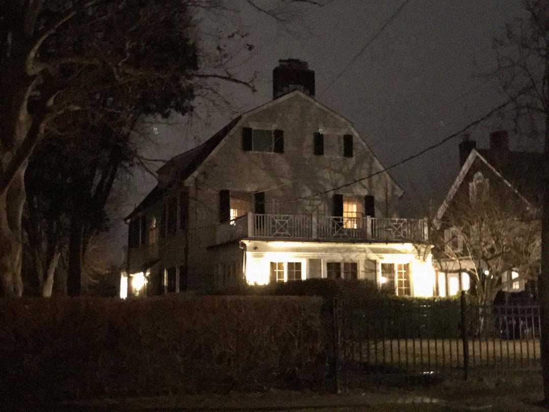 A large, two-story house with shuttered windows and a front porch is dimly lit at night, with warm lights illuminating the entrance and some windows. Bare trees and hedges surround the property.