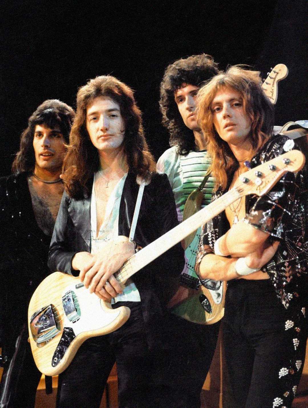 Four men with long hair stand together, holding musical instruments including a guitar and bass. They wear stylish, 1970s-style clothing and look directly at the camera against a dark background.