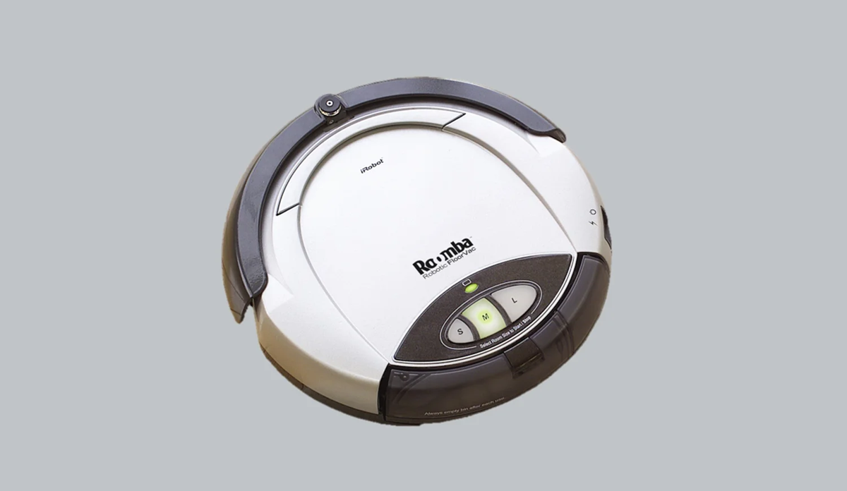 A white and gray robotic vacuum cleaner, labeled "Roomba," is shown on a plain gray background. The device has control buttons and indicator lights on its top surface.