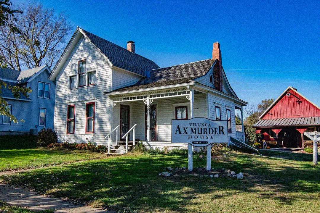 A white two-story house with a covered front porch stands next to a sign reading "Villisca Ax Murder House." A red barn is visible in the background under a clear blue sky.