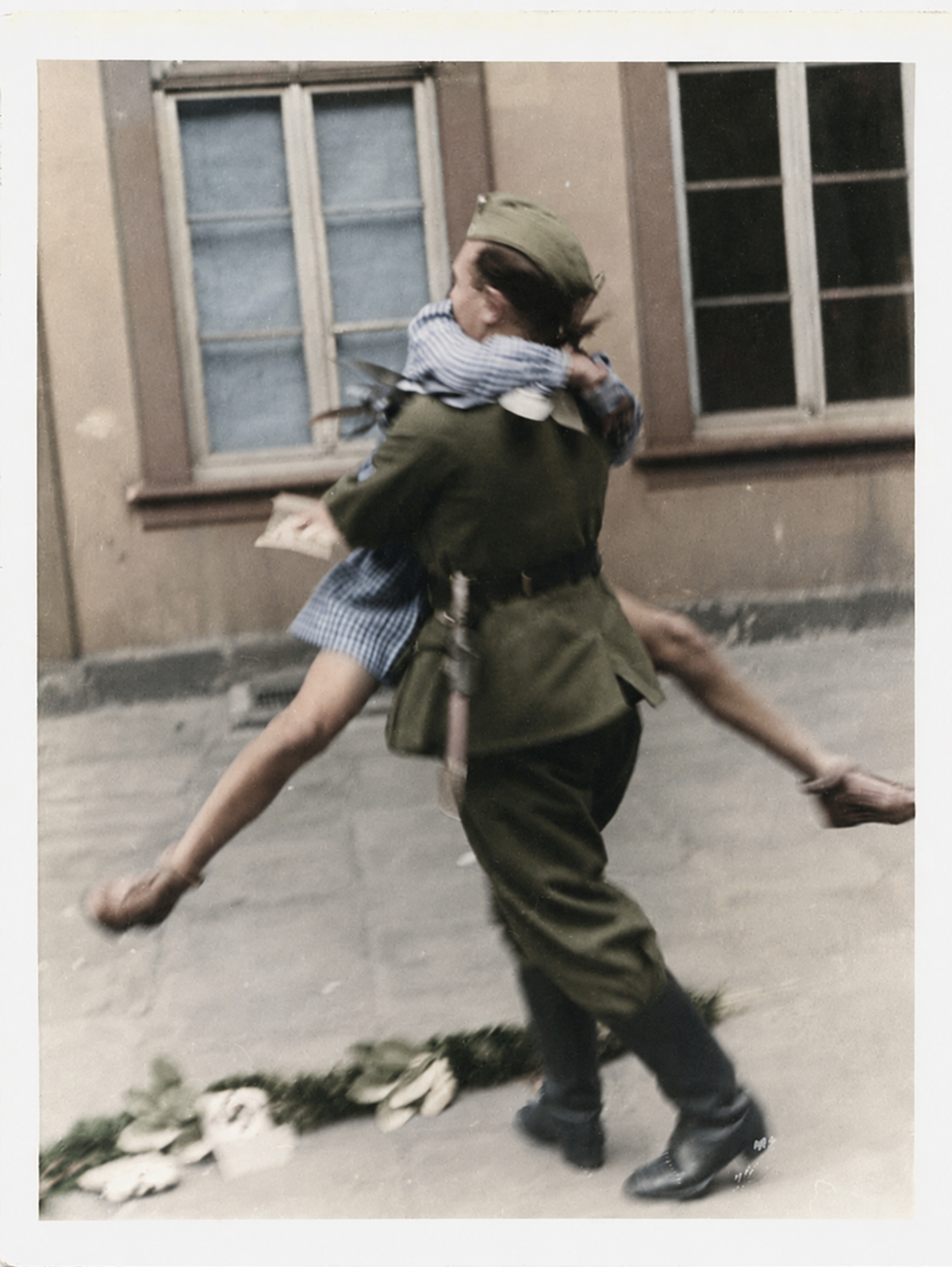 A soldier in uniform carries a child dressed in a blue checkered dress along a city street, passing by a building with two windows. The child's arms are around the soldier's neck.
