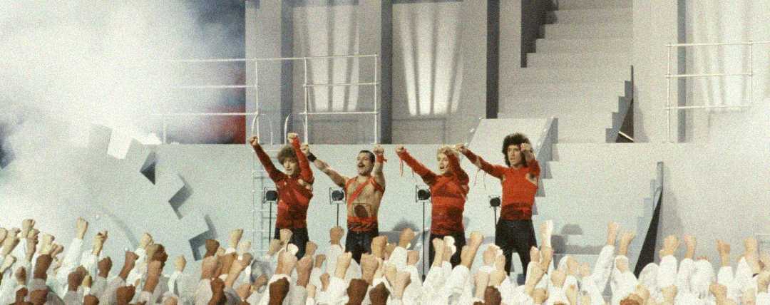 Three performers in red costumes stand on a white stage with geometric designs, raising their arms in front of a large crowd dressed in white, also raising their fists. The scene is dramatic and theatrical with smoke effects.