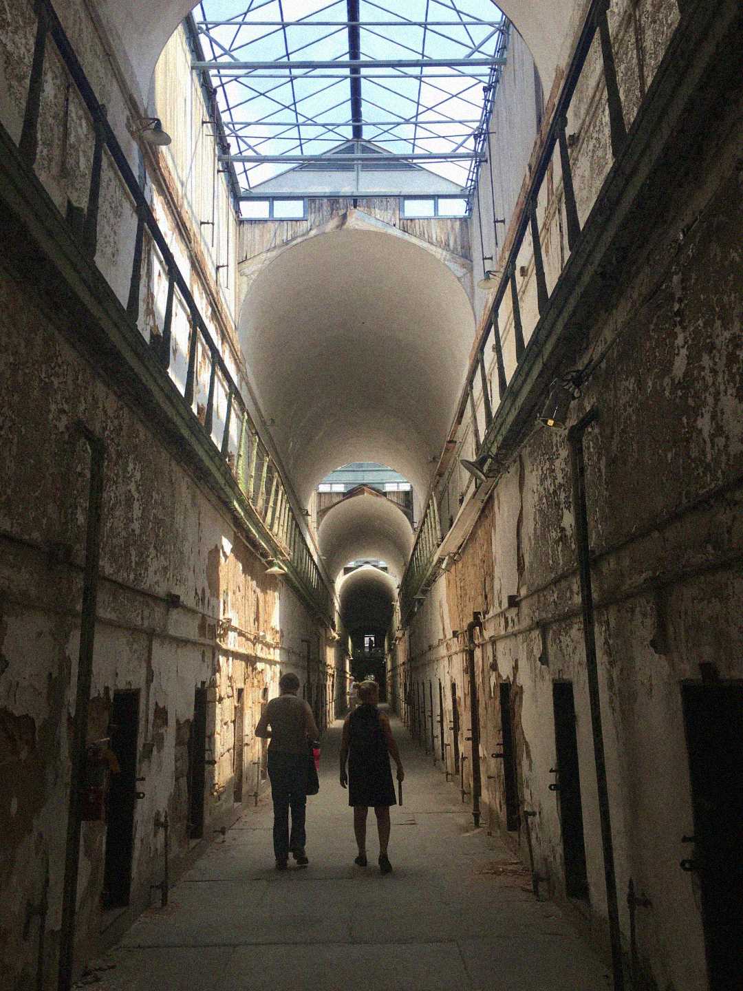 Two people walk down a long, narrow hallway with high arched ceilings and peeling walls, flanked by doors on both sides, under a partially glass roof letting in sunlight.