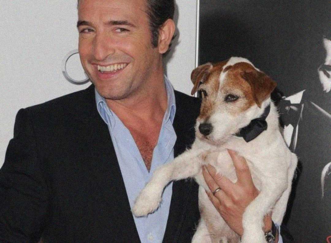 A smiling man in a suit holds a small white and brown dog wearing a black bow tie. They are standing in front of a black and white poster.