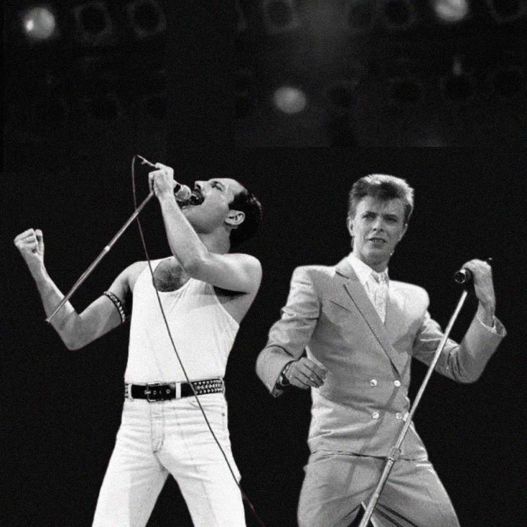 Two male singers perform energetically on stage; one wears a white tank top and pants, singing passionately into a microphone, while the other in a light-colored suit holds a microphone stand. The background is dark.