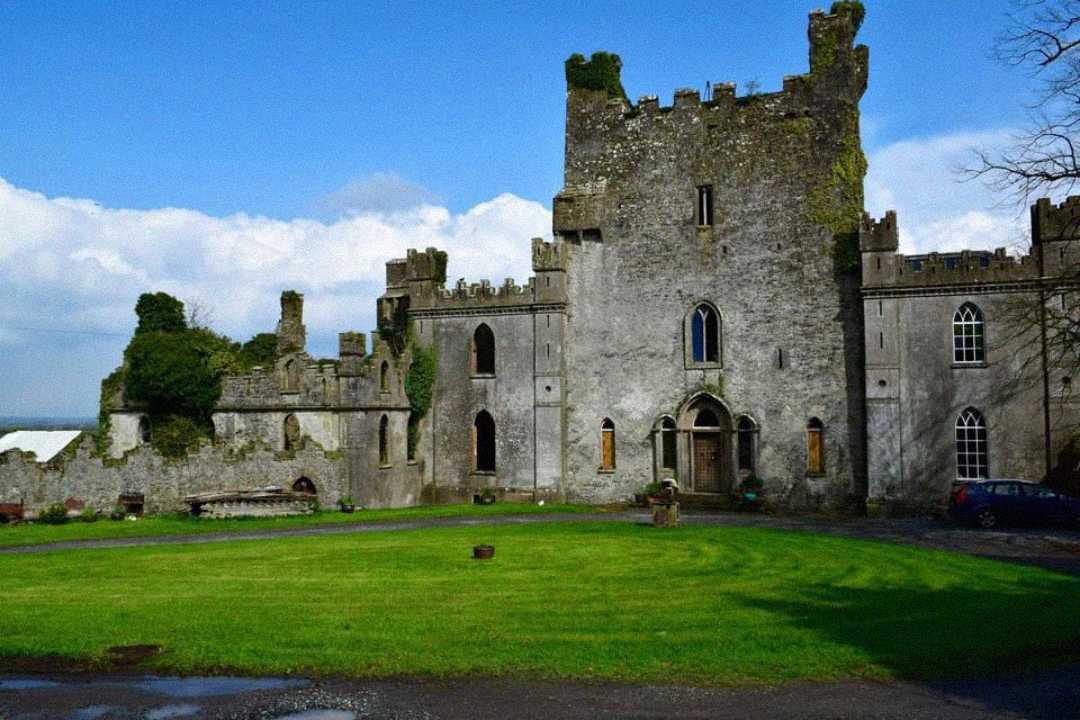 A large stone castle with ivy-covered walls, arched windows, and a turret, surrounded by a green lawn under a blue sky with scattered clouds.