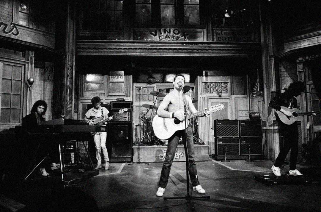 A black-and-white photo of a five-member band performing on stage, with a lead singer playing guitar at the center, flanked by musicians on keyboard, electric guitar, bass, and drums in a rustic, studio-like setting.