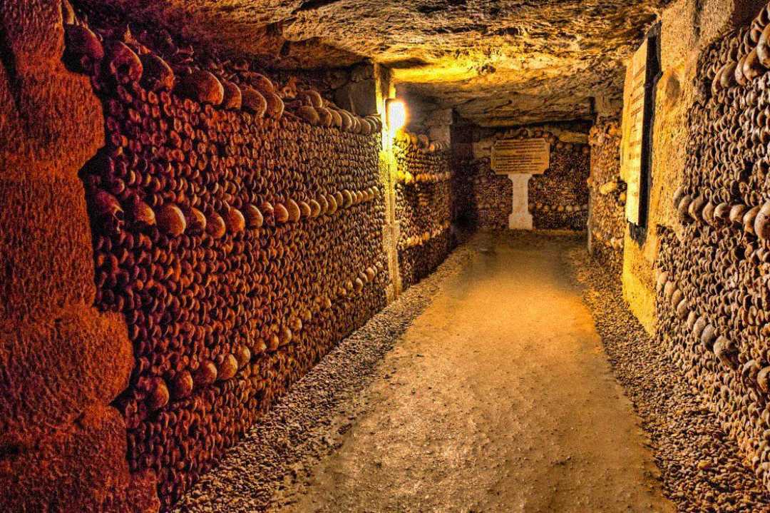 A dimly lit underground tunnel lined with human skulls and bones stacked neatly along the walls; a sign is visible at the end of the corridor, giving the scene an eerie and historic atmosphere.