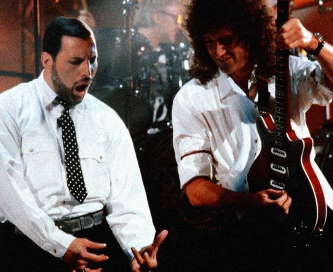 Two musicians perform on stage; one passionately sings in a white shirt and polka-dot tie, while the other, also in white, plays an electric guitar. Drums and stage lights are visible in the background.