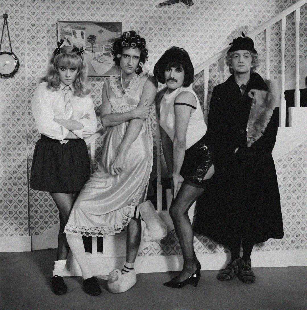 Four people pose in a vintage living room; each is dressed in humorous and exaggerated costume outfits, including robes, skirts, wigs, and accessories, imitating playful and theatrical characters.