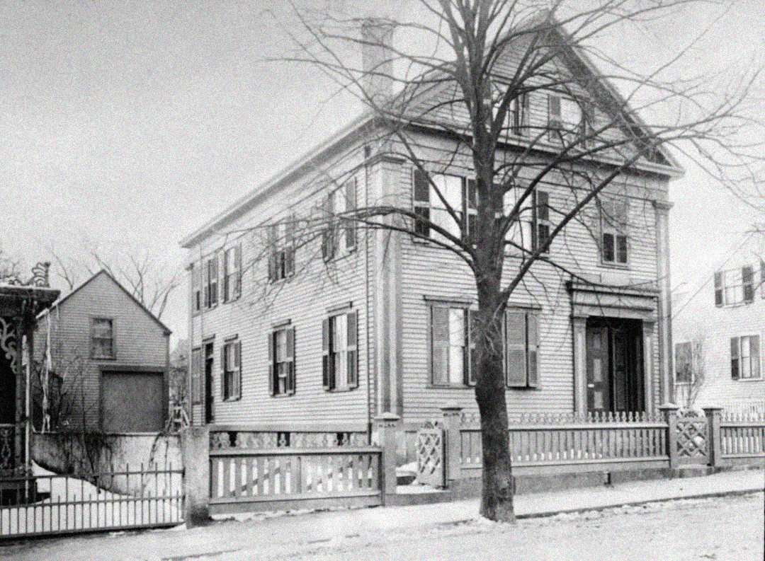 A black and white photograph of a two-story wooden house with shutters, a front porch, a bare tree in the yard, and a wooden fence along the sidewalk. Snow is visible on the ground and rooftops.