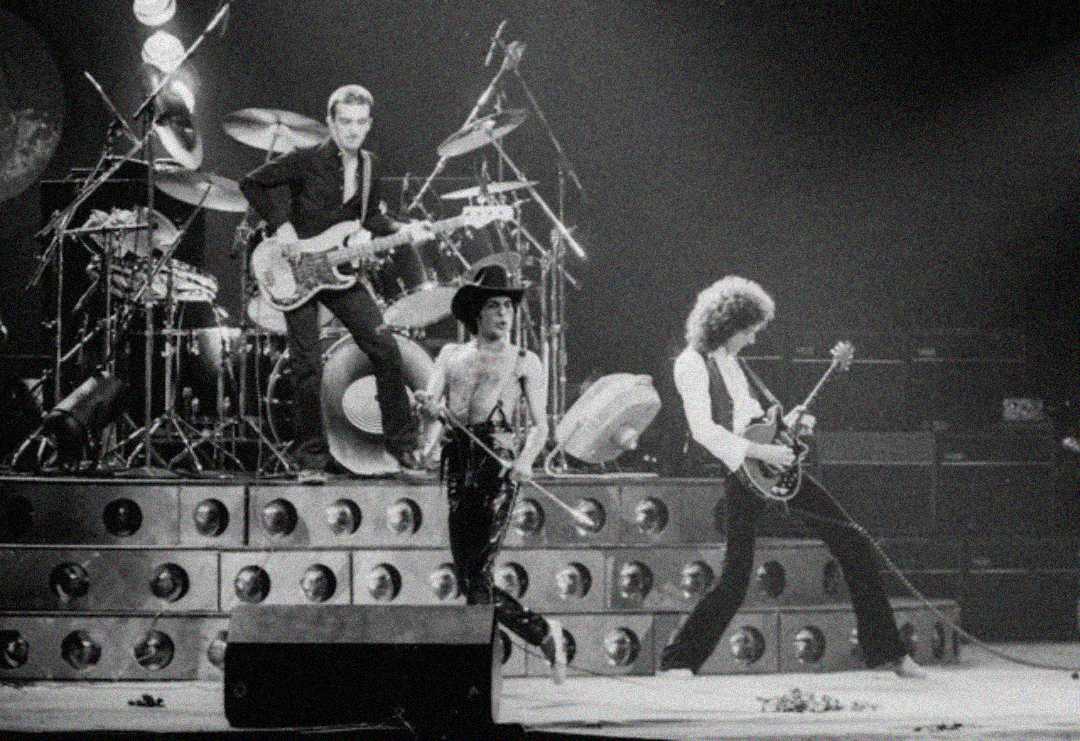 A black-and-white photo of a rock band performing on stage with dynamic lighting. One member plays bass, another sings and dances at the center, and two others are on guitar and drums. Instruments and stage equipment are visible.
