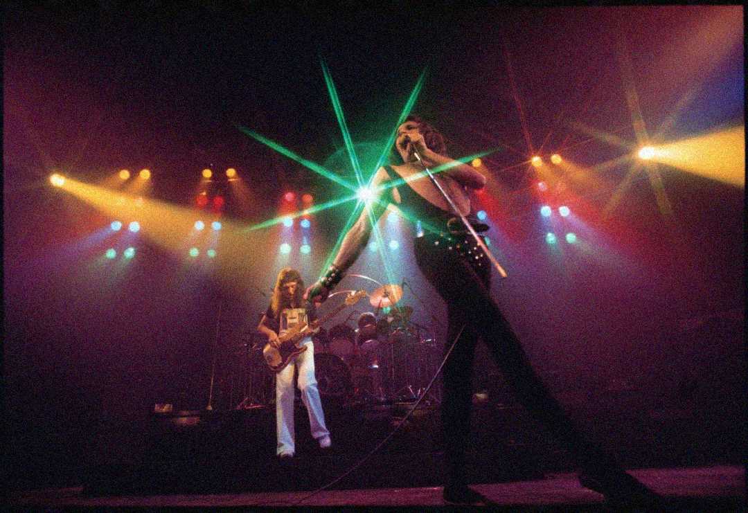 A dynamic rock concert scene with two musicians performing on stage under colorful, dramatic lighting; one sings passionately into a microphone while the other plays guitar in the background.