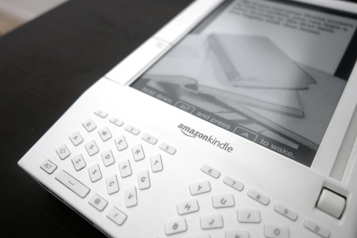 Close-up of an early model Amazon Kindle e-reader with a physical keyboard, displaying an e-ink screen showing an image of stacked books. The device is placed on a dark surface.
