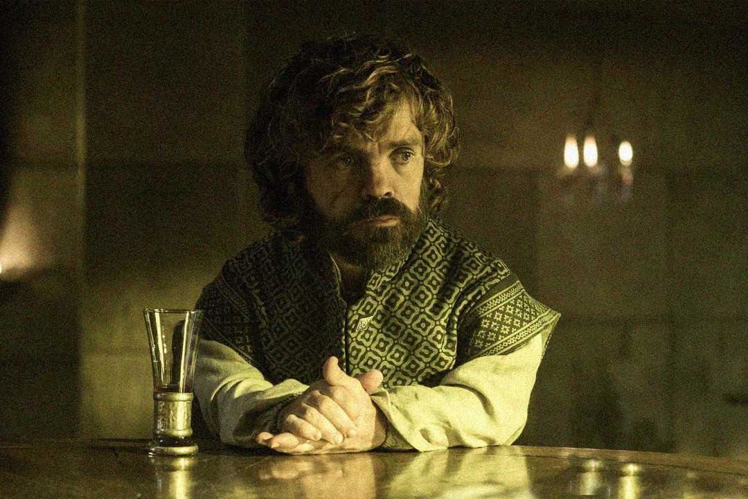 A man with curly hair and a beard sits at a table with his hands clasped, wearing patterned robes. A glass sits nearby, and the dim, warm lighting suggests a medieval or fantasy setting.