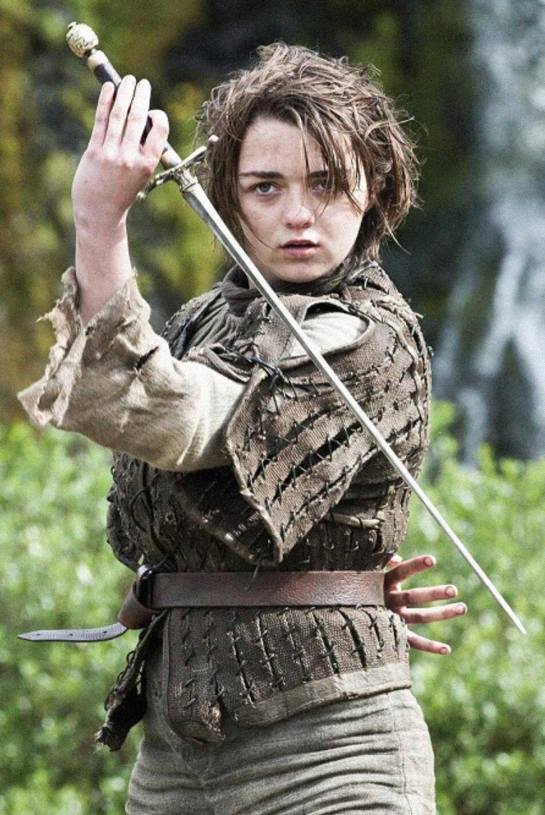 A young person with short, messy hair and worn, medieval-style clothing holds a sword in a defensive stance outdoors, with greenery and a blurred background behind them.