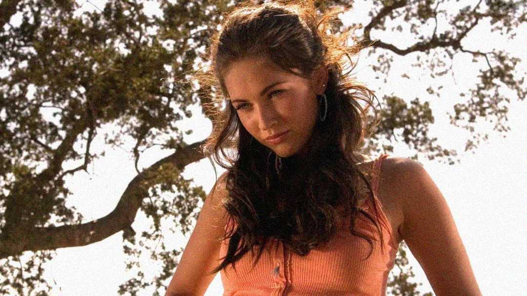 A young woman with long brown hair stands outdoors, wearing a peach-colored sleeveless top. Sunlight filters through the trees behind her, creating a warm, natural background.