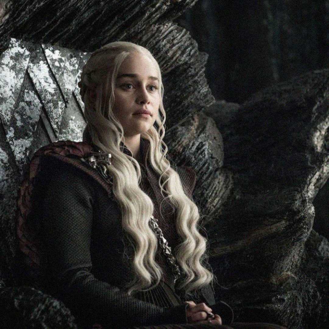 A woman with long, pale blonde hair and a dark, intricate outfit sits on a dark, stone throne in a dimly lit room, looking serious and thoughtful.