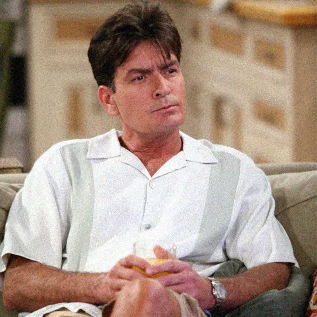 A man with dark hair wearing a short-sleeved, white and light gray shirt sits on a couch holding a drink, looking to the side with a thoughtful expression. The background shows light-colored furniture.