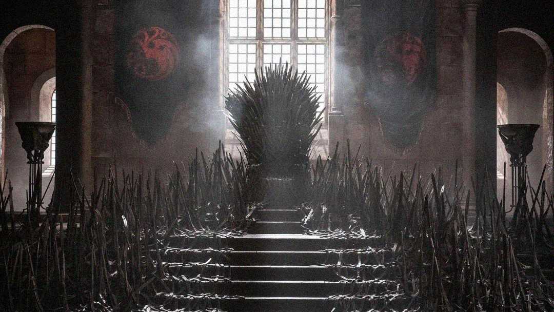 A dramatic, dimly lit room features an imposing iron throne made of swords at the top of stone steps, flanked by two dragon banners, with light streaming in through large windows behind it.