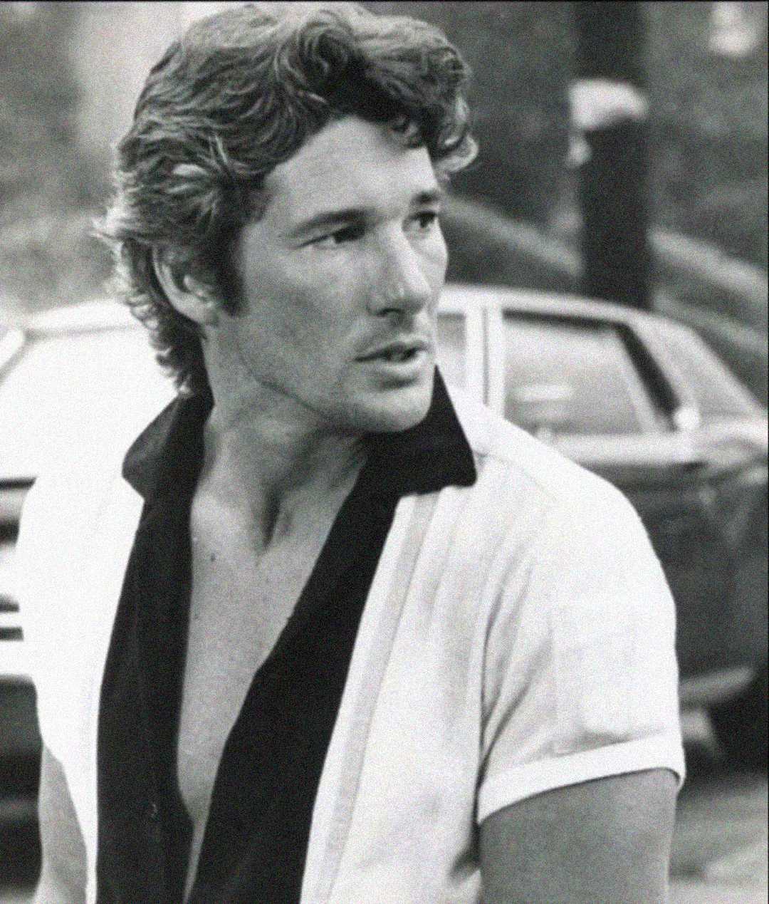 A man with wavy hair wearing a partially unbuttoned short-sleeve shirt looks to the side. The image is black and white, and there is a car in the blurred background.