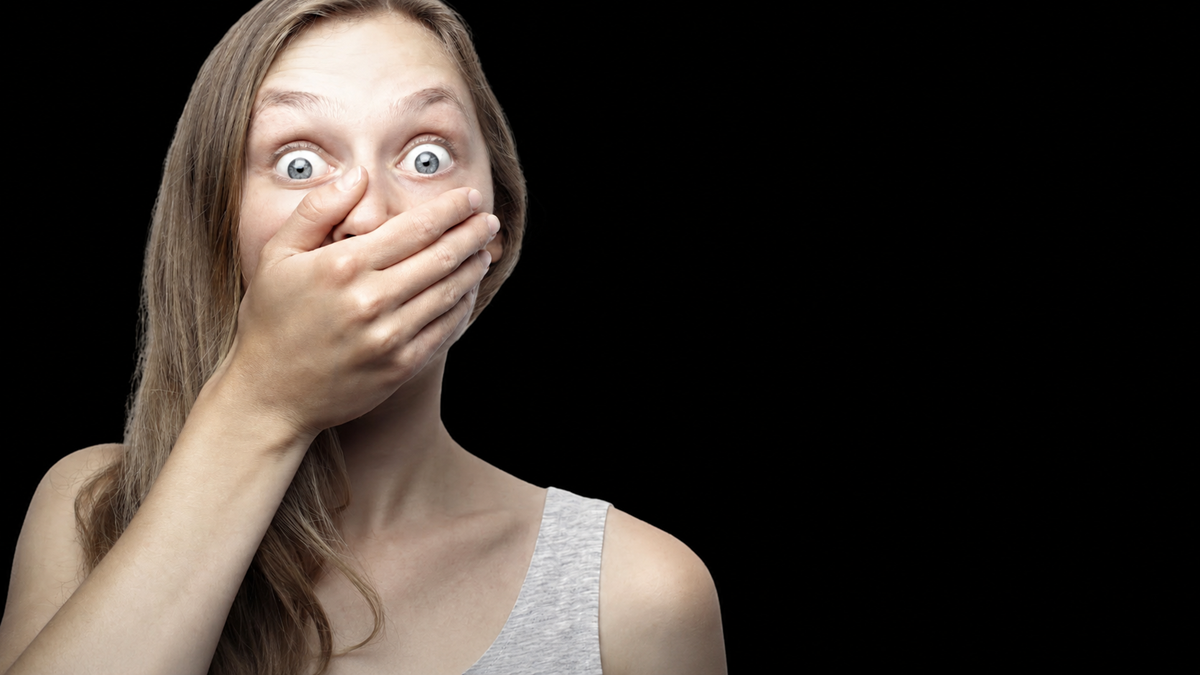 A young woman with long hair and wide eyes covers her mouth with her hand, appearing shocked or surprised, against a plain black background.