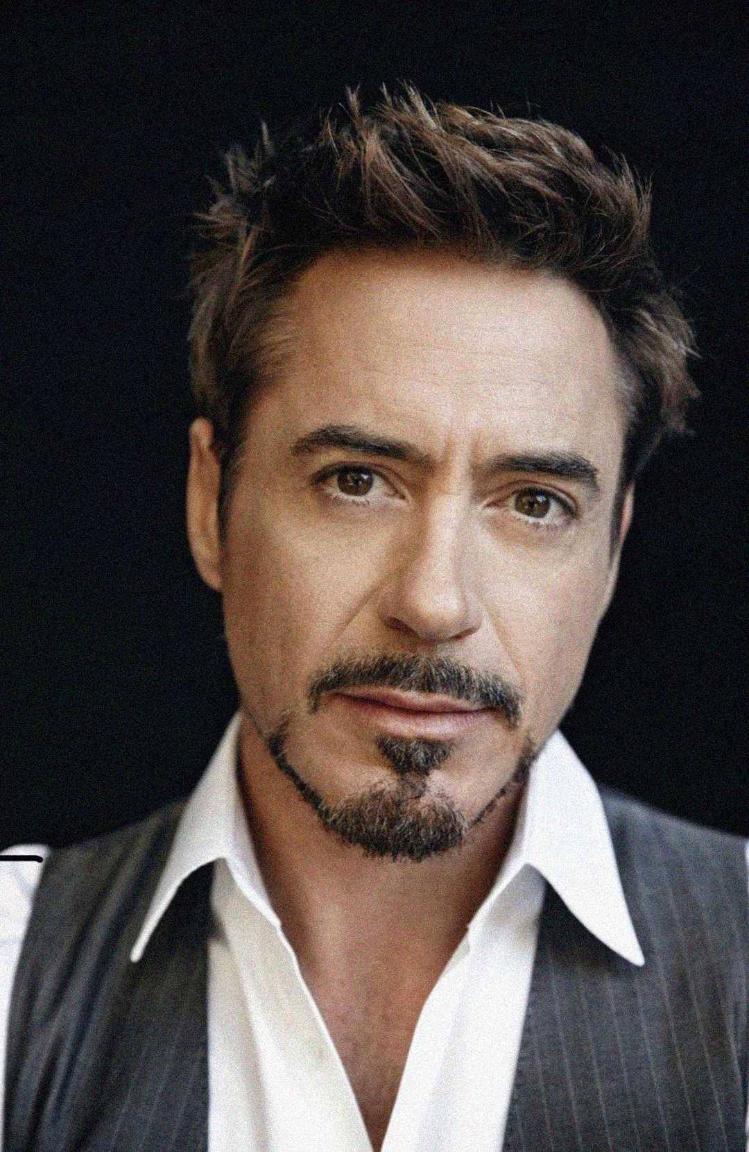 A man with short, styled brown hair and a goatee wearing a white collared shirt and gray vest looks directly at the camera against a dark background.