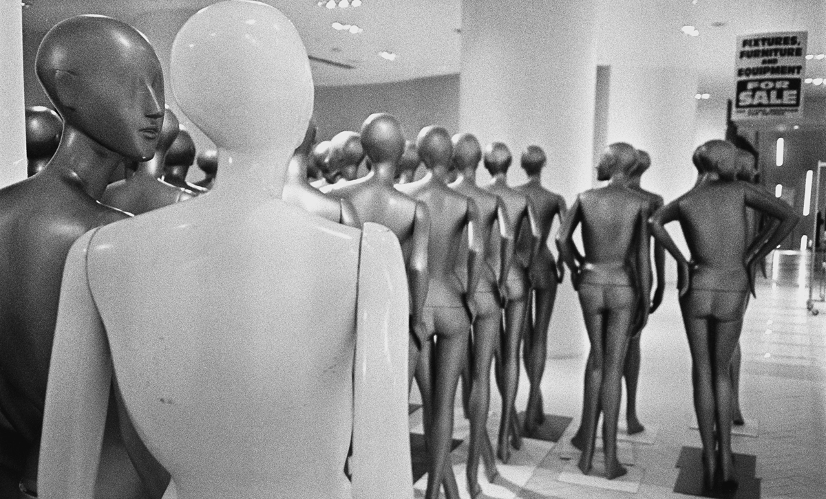 A group of mannequins are arranged in rows inside a store, facing away from the camera. The black-and-white image shows a “For Sale” sign in the background advertising fixtures, furniture, and equipment.