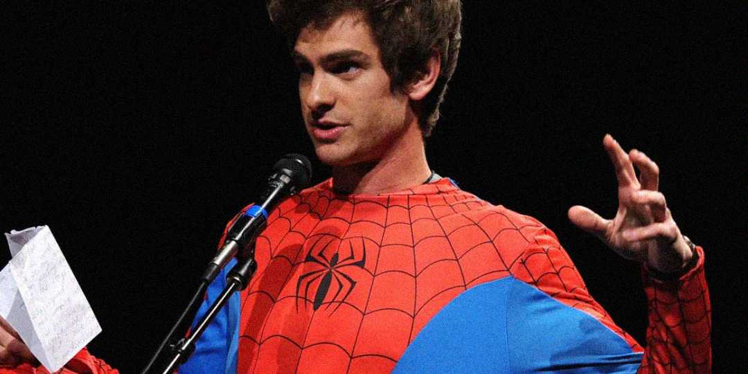 A person wearing a red and blue Spider-Man costume speaks into a microphone while holding a piece of paper, gesturing with their other hand against a dark background.