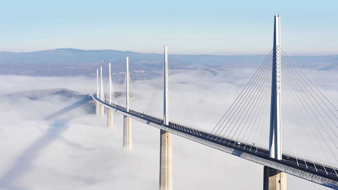 A long cable-stayed bridge with tall towers rises above thick clouds, stretching across a fog-covered valley, with distant hills visible in the background.