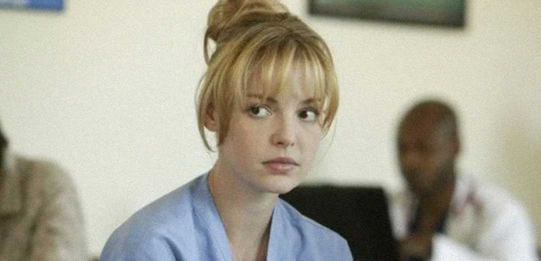 A young woman with blonde hair in a loose bun, wearing a light blue top, looks slightly to the side with a neutral expression. The background is softly blurred with a person sitting behind her.