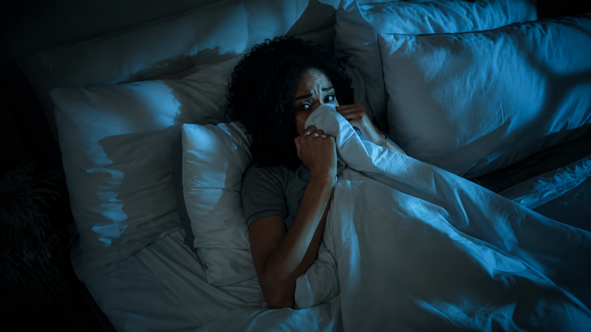 A woman lies in bed at night, looking scared and clutching a blanket up to her face. The room is dimly lit with blue light, and her wide eyes suggest fear or anxiety.