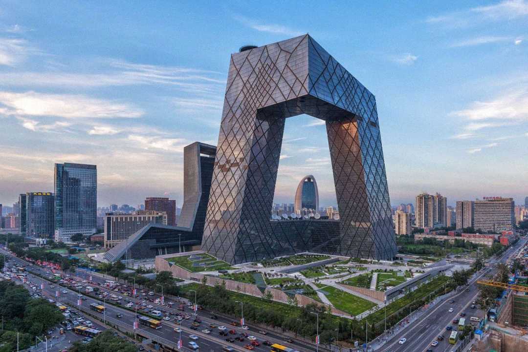 A large, modern skyscraper with a unique looped design stands in an urban cityscape, surrounded by smaller buildings, green spaces, and busy roads, under a partly cloudy sky.