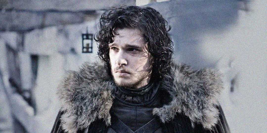 A man with curly dark hair wearing a fur-lined cloak stands outdoors in a snowy, stone setting, looking serious and slightly to the side.