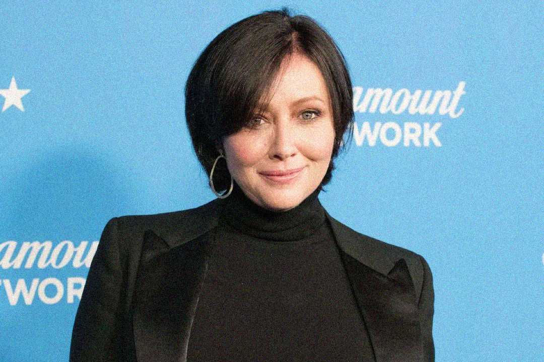 A woman with short dark hair, wearing a black turtleneck and black blazer, stands in front of a blue background with white text that reads "Paramount Network." She is smiling slightly.