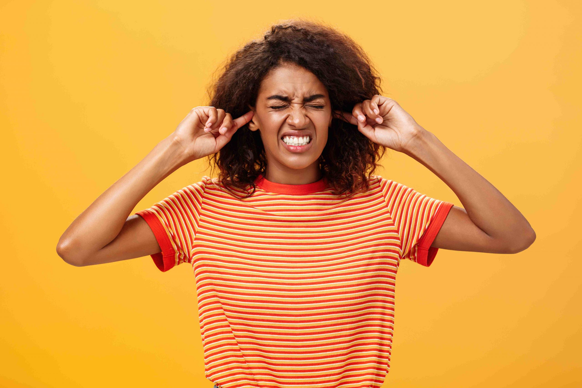 A young woman in a red and white striped shirt stands against an orange background, closing her eyes and plugging her ears with her fingers, appearing annoyed or frustrated by a loud noise.
